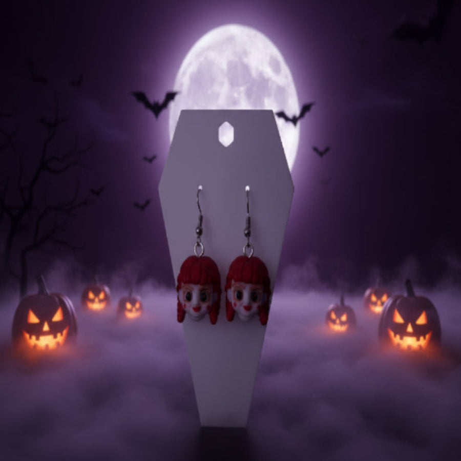 A pair of dangling earrings with small character faces featuring bright pink hair and white skin, displayed on a white coffin-shaped stand on a wooden desk with a blurred keyboard and room in the background.