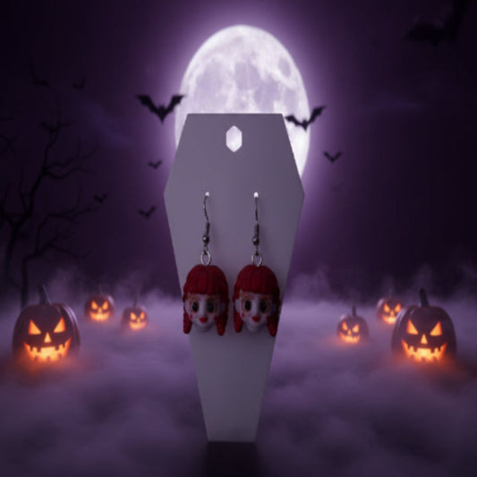 A pair of dangling earrings with small character faces featuring bright pink hair and white skin, displayed on a white coffin-shaped stand on a wooden desk with a blurred keyboard and room in the background.